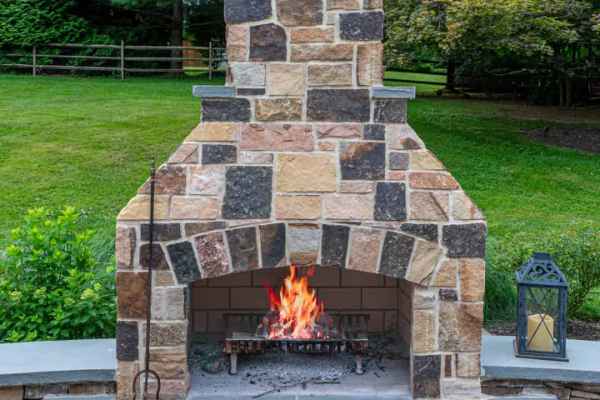 Stone outdoor fireplace with a burning fire sits amidst a beautiful slate patio, crafted with expert hardscaping services in Delaware County, PA. A small lantern and fireplace tools rest on the stone ledge, while lush green grass and trees provide a serene backdrop for your perfect outdoor living experience.