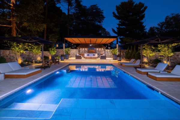 Luxurious outdoor pool area at dusk with soft lighting. Features four sun loungers, two large umbrellas, and a stylish seating area under a modern pergola. Expert hardscaping services in Delaware County, PA enhance the serene atmosphere with trees and stone walls in the background.