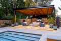 Modern outdoor living space with a partially covered pergola, featuring a cozy seating area, TV, and ceiling fan. Surrounded by large potted plants and crafted through premium hardscaping services in Delaware County, PA, it overlooks a rectangular pool. Stone walls and greenery enhance the serene atmosphere.