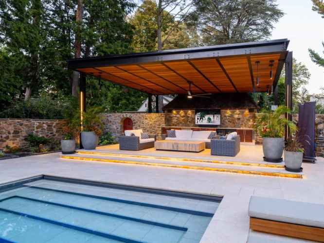 Modern outdoor living space with a partially covered pergola, featuring a cozy seating area, TV, and ceiling fan. Surrounded by large potted plants and crafted through premium hardscaping services in Delaware County, PA, it overlooks a rectangular pool. Stone walls and greenery enhance the serene atmosphere.