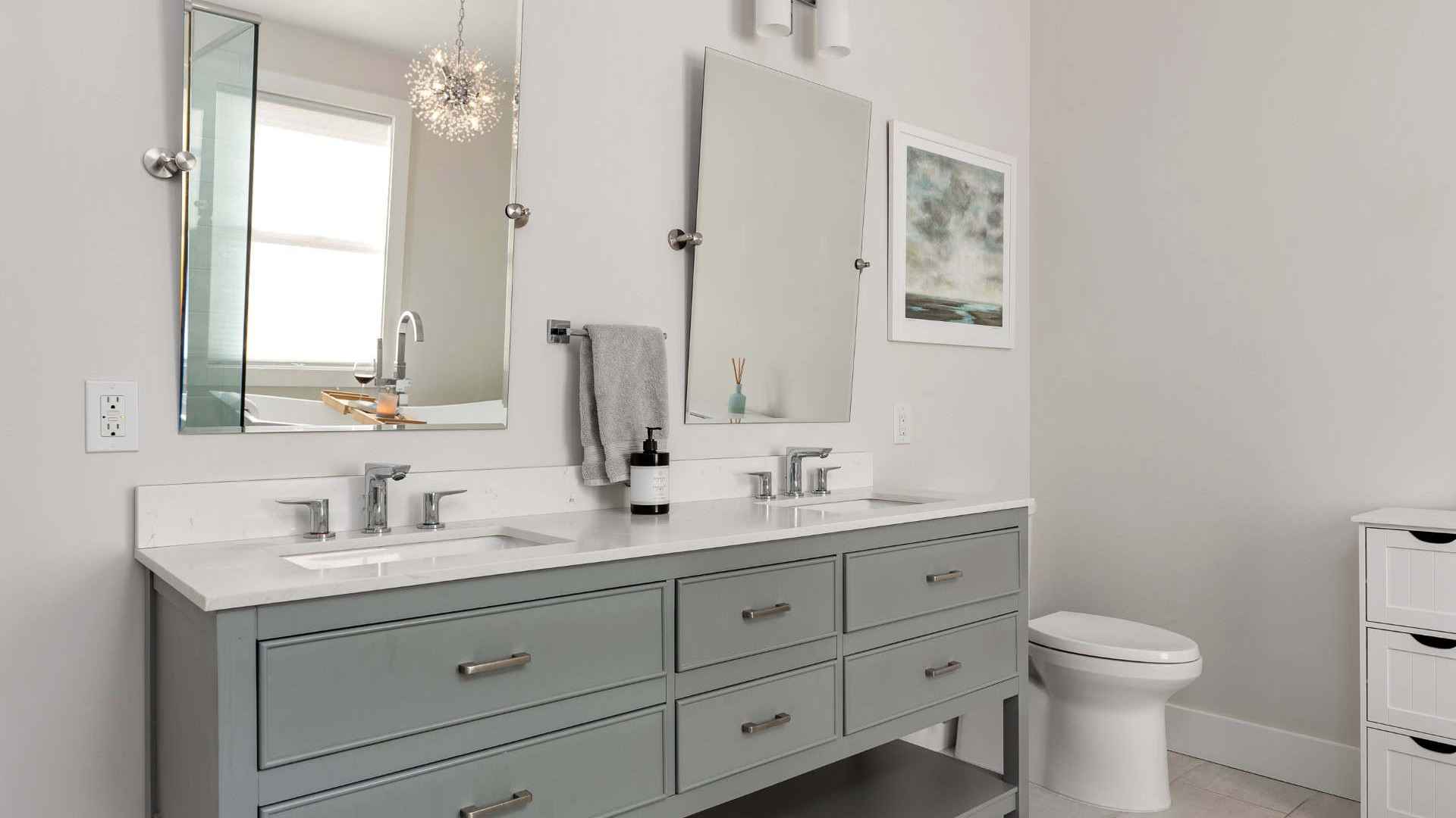 Modern bathroom with a double vanity featuring gray cabinets and two mirrors above. A white countertop with dual sinks and a sleek soap dispenser completes the look. To the right, find the toilet near a window adorned with blinds and wall art, echoing outdoor living vibes from Delaware County, PA.