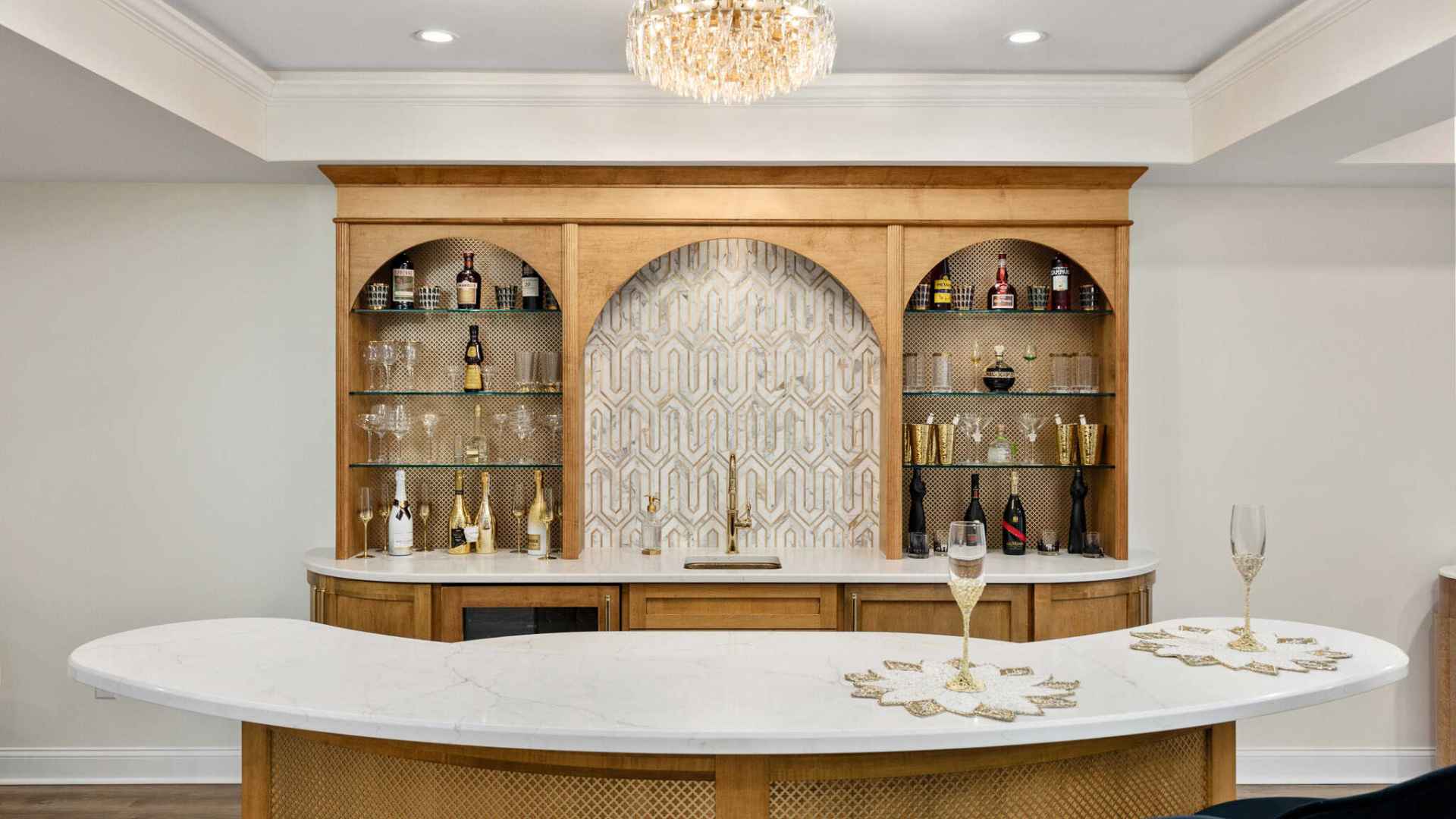 A stylish home bar with a curved white countertop and gold accents. Wooden shelves with decorative bottles flank an ornate patterned backsplash. A crystal chandelier hangs above, illuminating the space elegantly—perfect for discussing outdoor living in Delaware County, PA over wine.