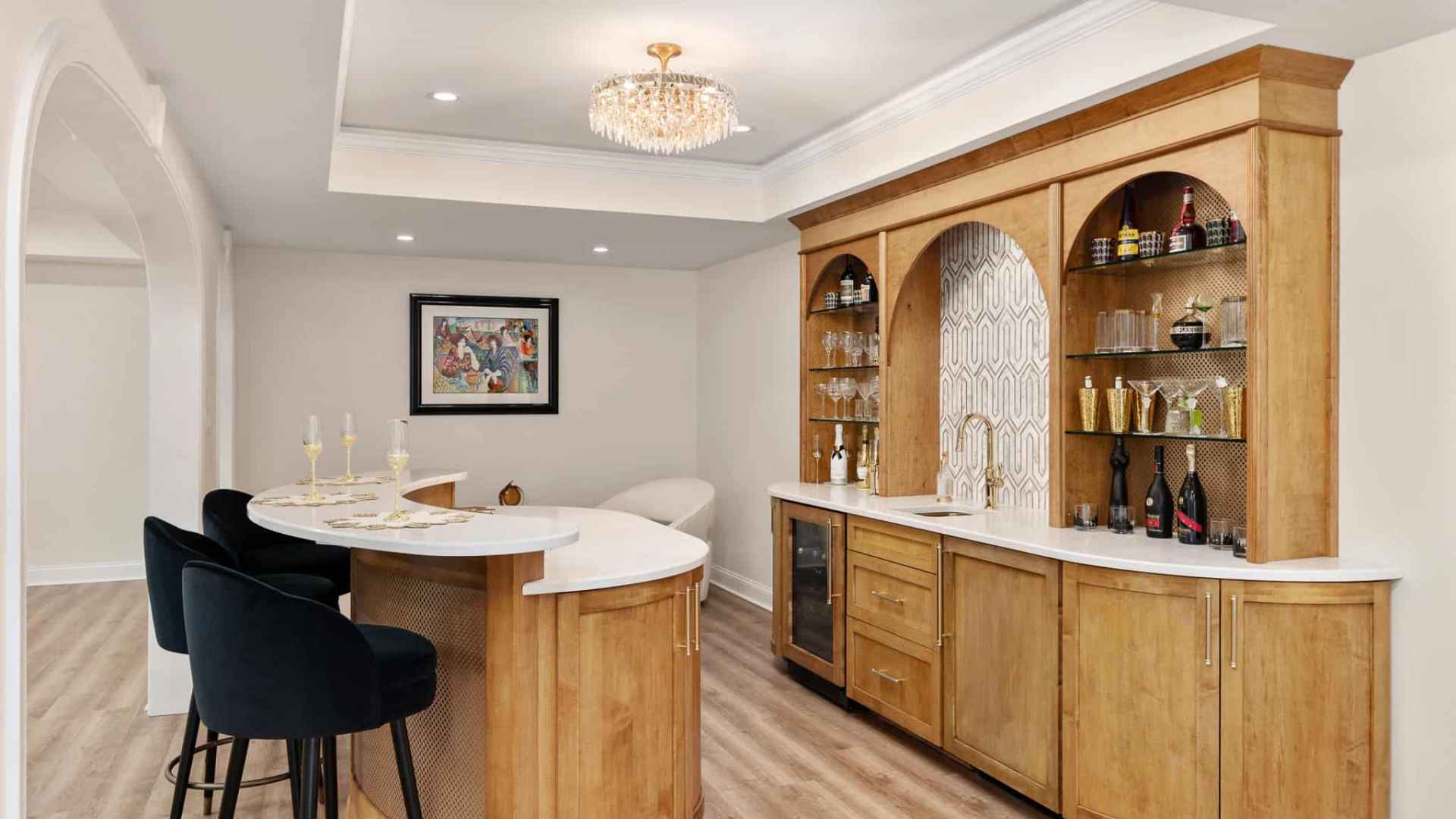A modern home bar with a curved white countertop, wooden cabinetry, and a decorative backsplash offers the perfect indoor complement to your outdoor living space in Delaware County, PA. Shelves display bottles and glassware while dark upholstered stools sit beneath a chandelier’s glow.