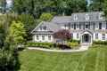 A large, two-story stone and siding house with white shutters and a grey roof sits amidst lush greenery in Delaware County, PA. The well-maintained front lawn is complemented by neatly trimmed hedges and trees, showcasing expert landscaping services. A curved path leads to the entrance.