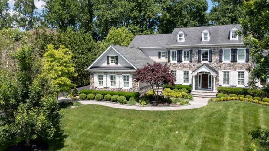 A large, two-story stone and siding house with white shutters and a grey roof sits amidst lush greenery in Delaware County, PA. The well-maintained front lawn is complemented by neatly trimmed hedges and trees, showcasing expert landscaping services. A curved path leads to the entrance.