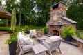 Outdoor living at its finest in Delaware County, PA, with a cozy stone fireplace surrounded by wooden chairs adorned with green and white cushions. A decorative centerpiece graces the table, while tall planters and lush greenery complete this serene setting.