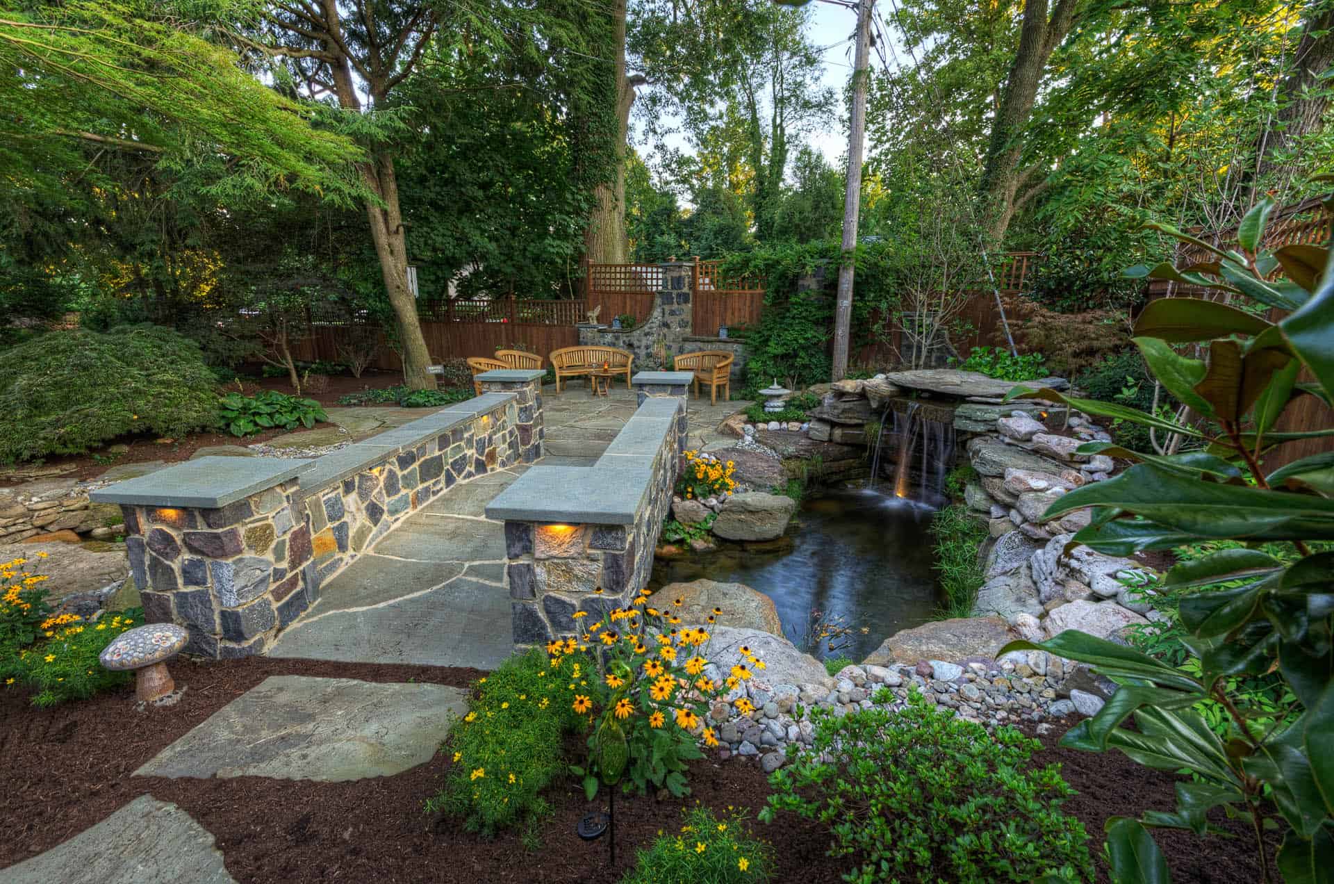 A landscaped backyard features a stone patio with seating, a small waterfall flowing into a pond, lush greenery, yellow flowers, and mature trees, creating a serene and inviting outdoor space.