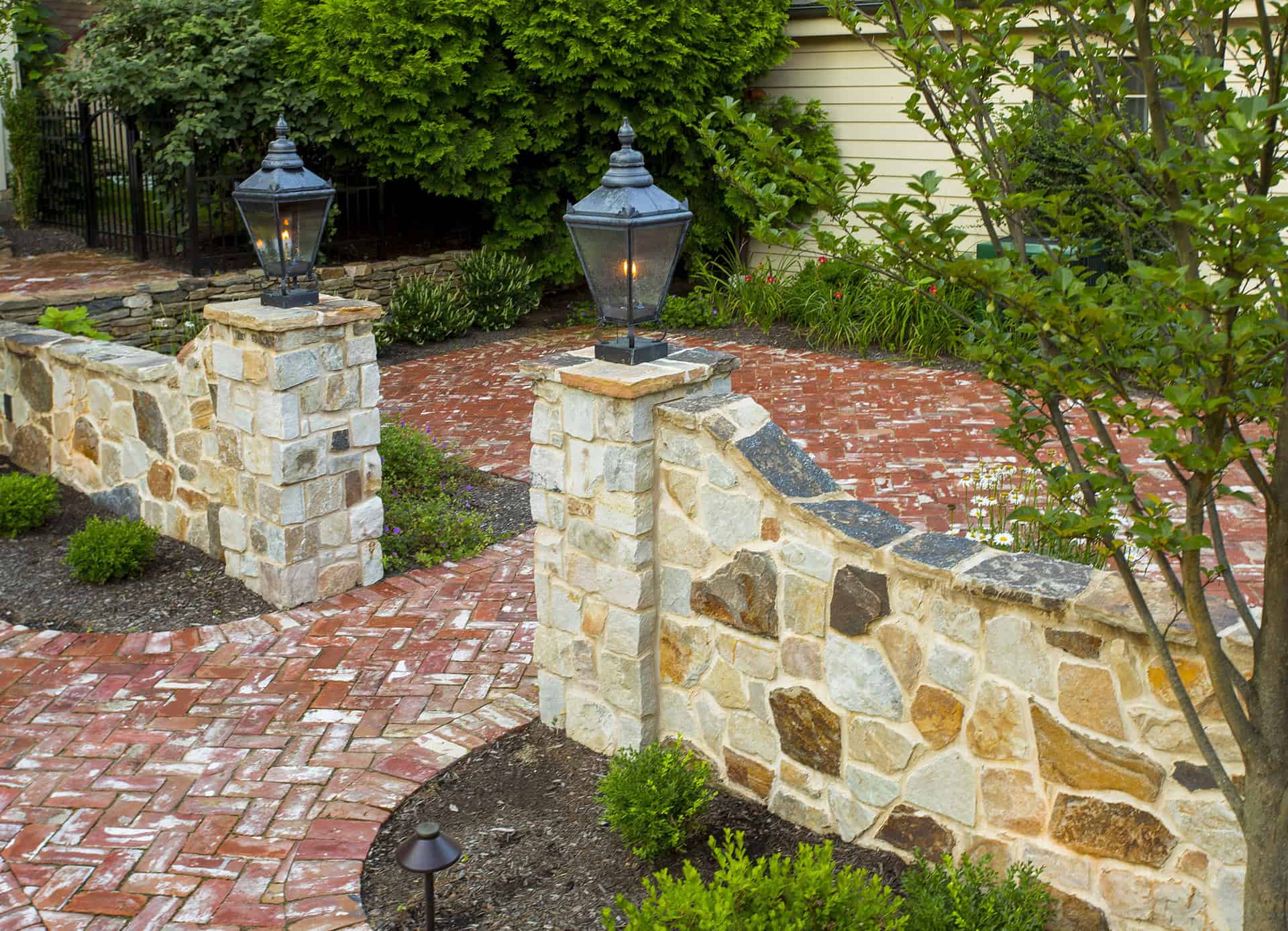 Curved stone wall with brick pillars topped by lanterns, bordering a red brick pathway and patio. Manicured green shrubs and trees line the garden area beside a yellow house.