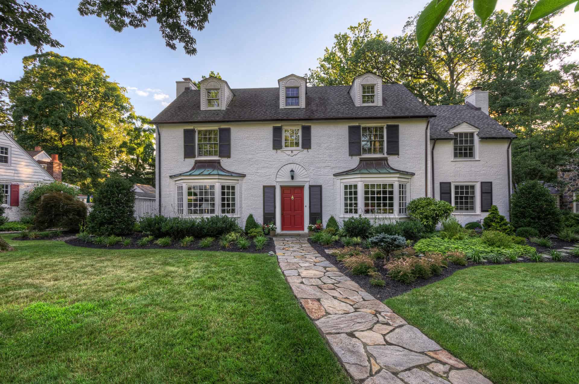 A large, two-story white house with a dark roof, red front door, and dormer windows. A stone walkway leads to the entrance, surrounded by manicured lawns, shrubs, and trees in the front yard.