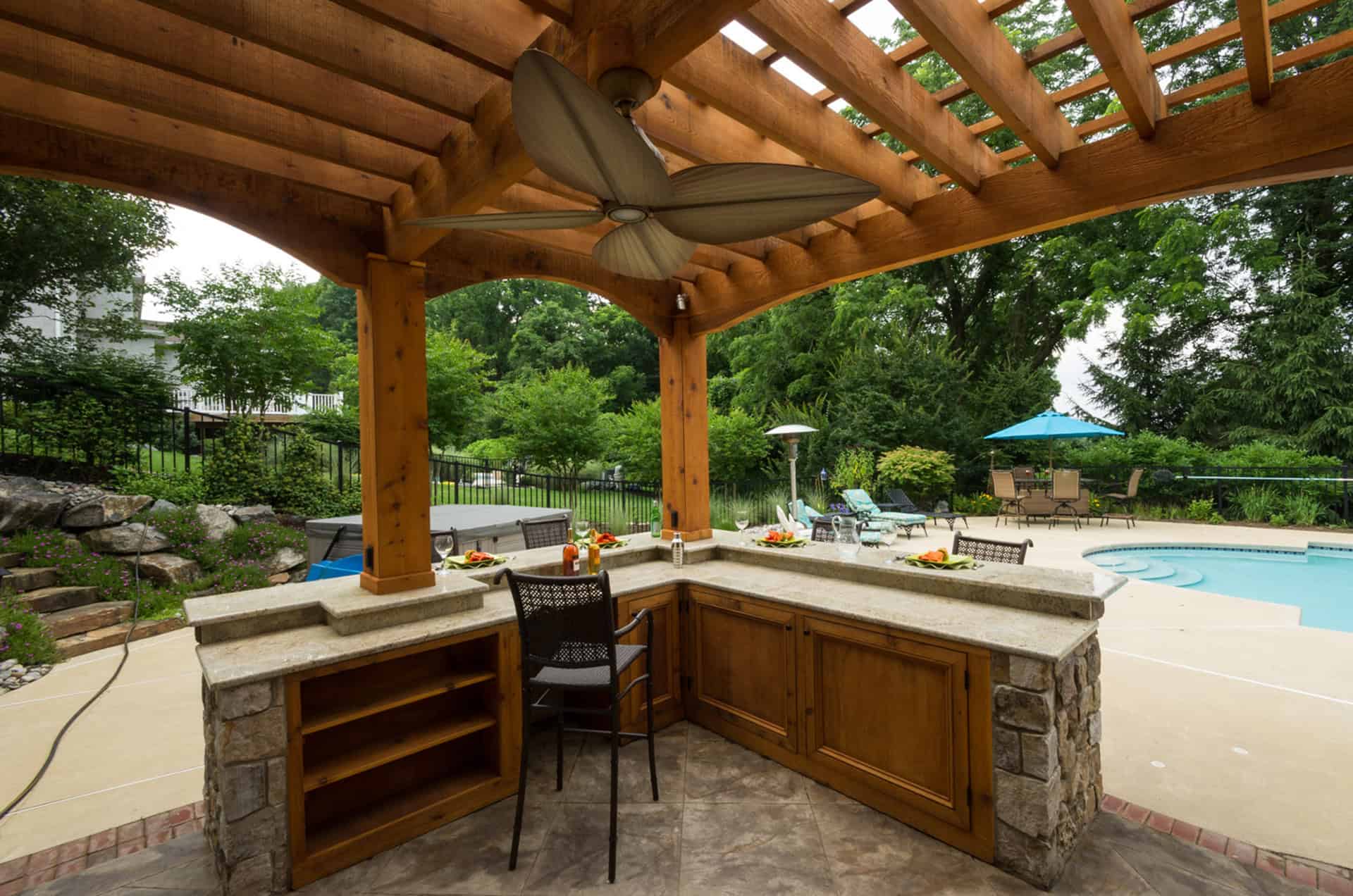 Outdoor kitchen with stone countertops and wooden pergola, two high chairs, and a ceiling fan, overlooking a swimming pool and patio area surrounded by green trees and landscaping.