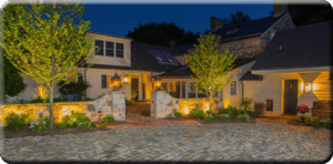 A charming house at night showcases exquisite stone and brick architecture, enhanced by warm exterior lighting. Flanking the entrance are two small trees, while a cobblestone path leads to a welcoming wooden door. Perfectly illustrating outdoor living in Delaware County, PA.