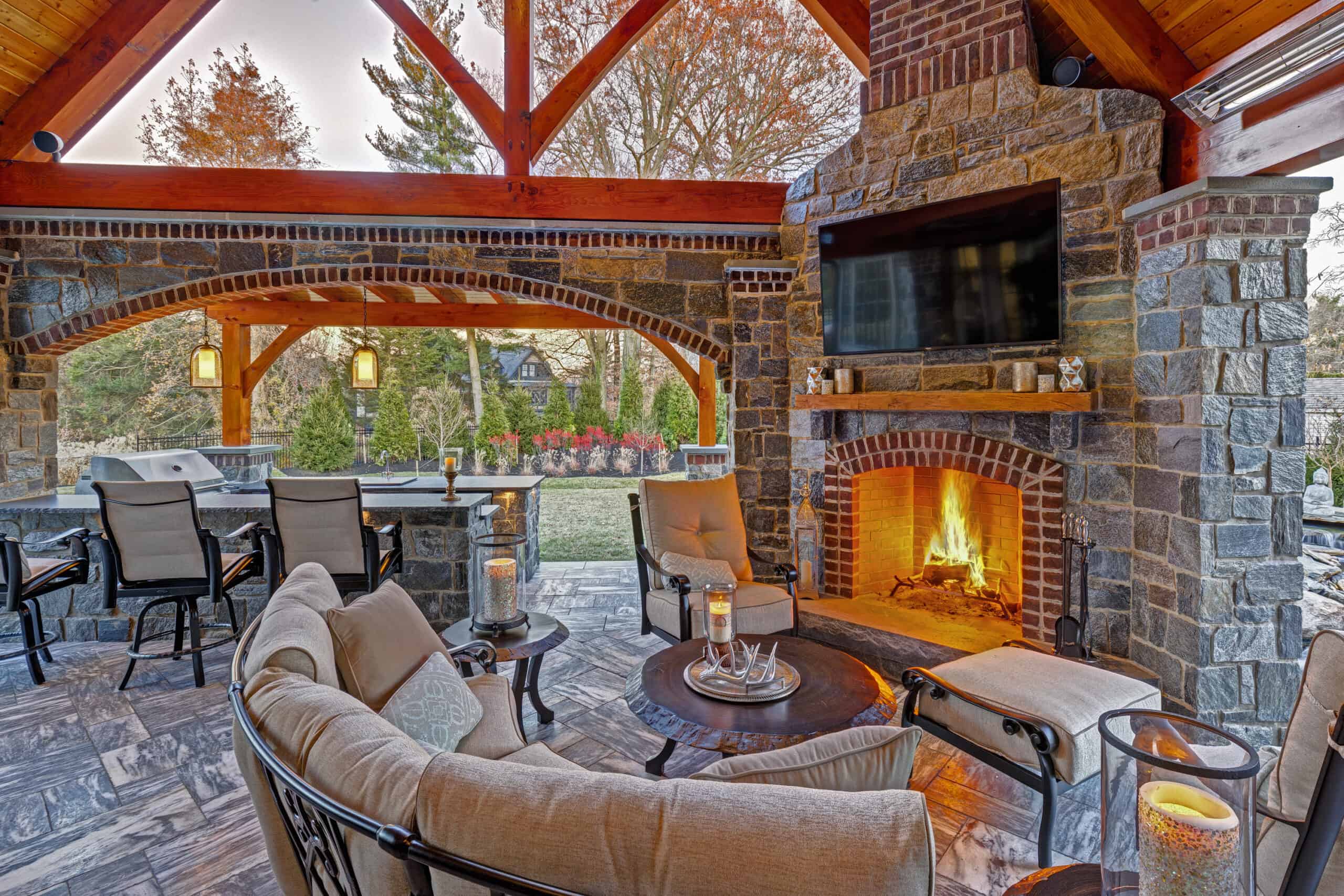 A cozy outdoor patio with cushioned seating around a fire table, a stone fireplace with a TV above, and a bar area with stools under a wooden pergola, all overlooking a landscaped garden.