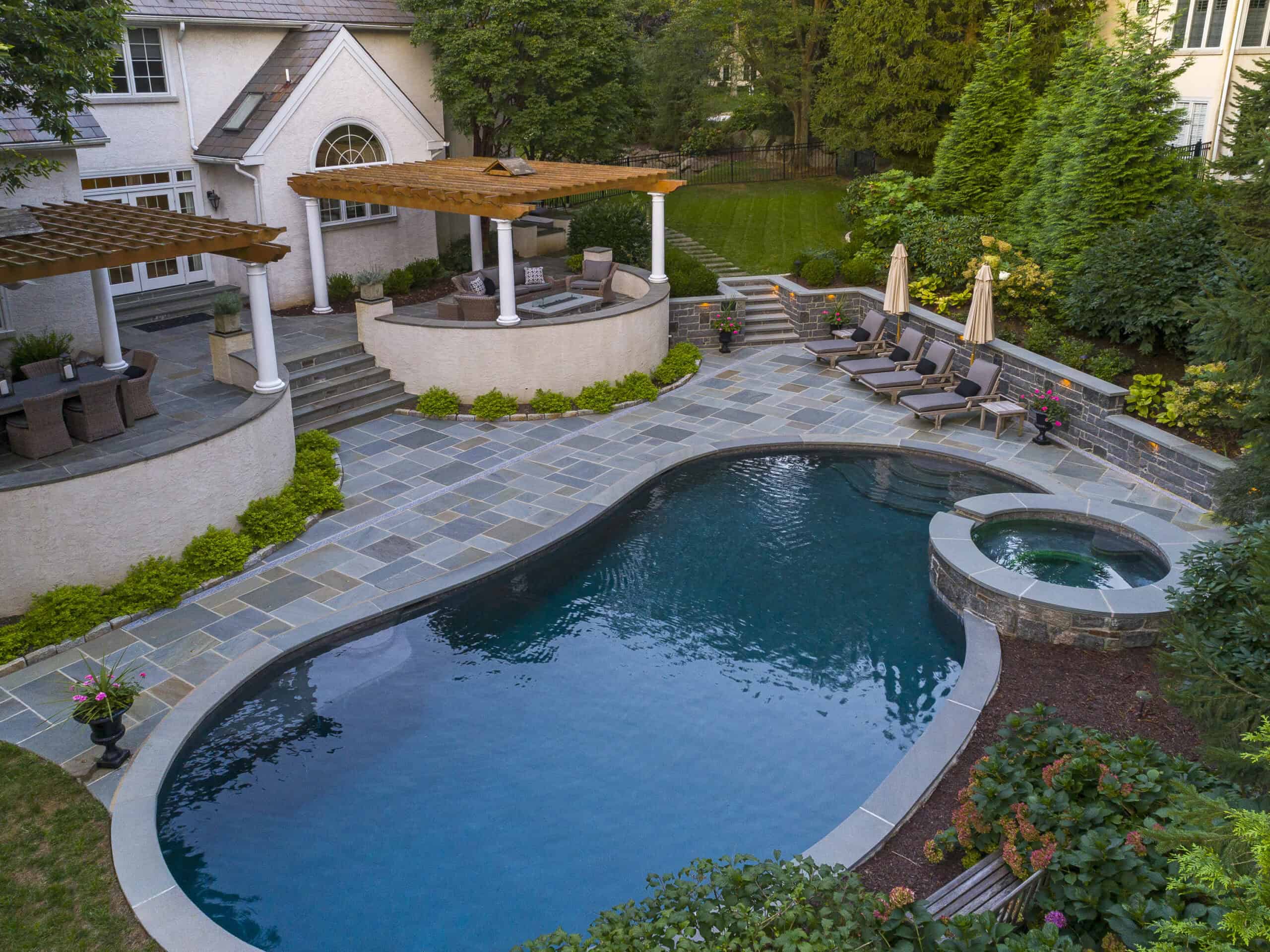 Backyard featuring a stunning kidney-shaped pool with an attached circular hot tub, stone patio, lounge chairs, umbrellas, pergolas, lush landscaped greenery, and a large house in the background.