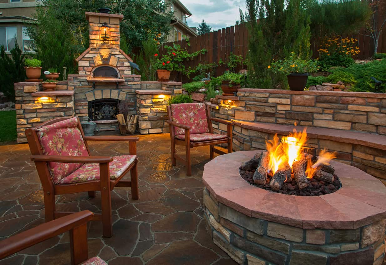 A backyard patio with a fire pit and stone fireplace, wooden chairs, and ambient lighting.