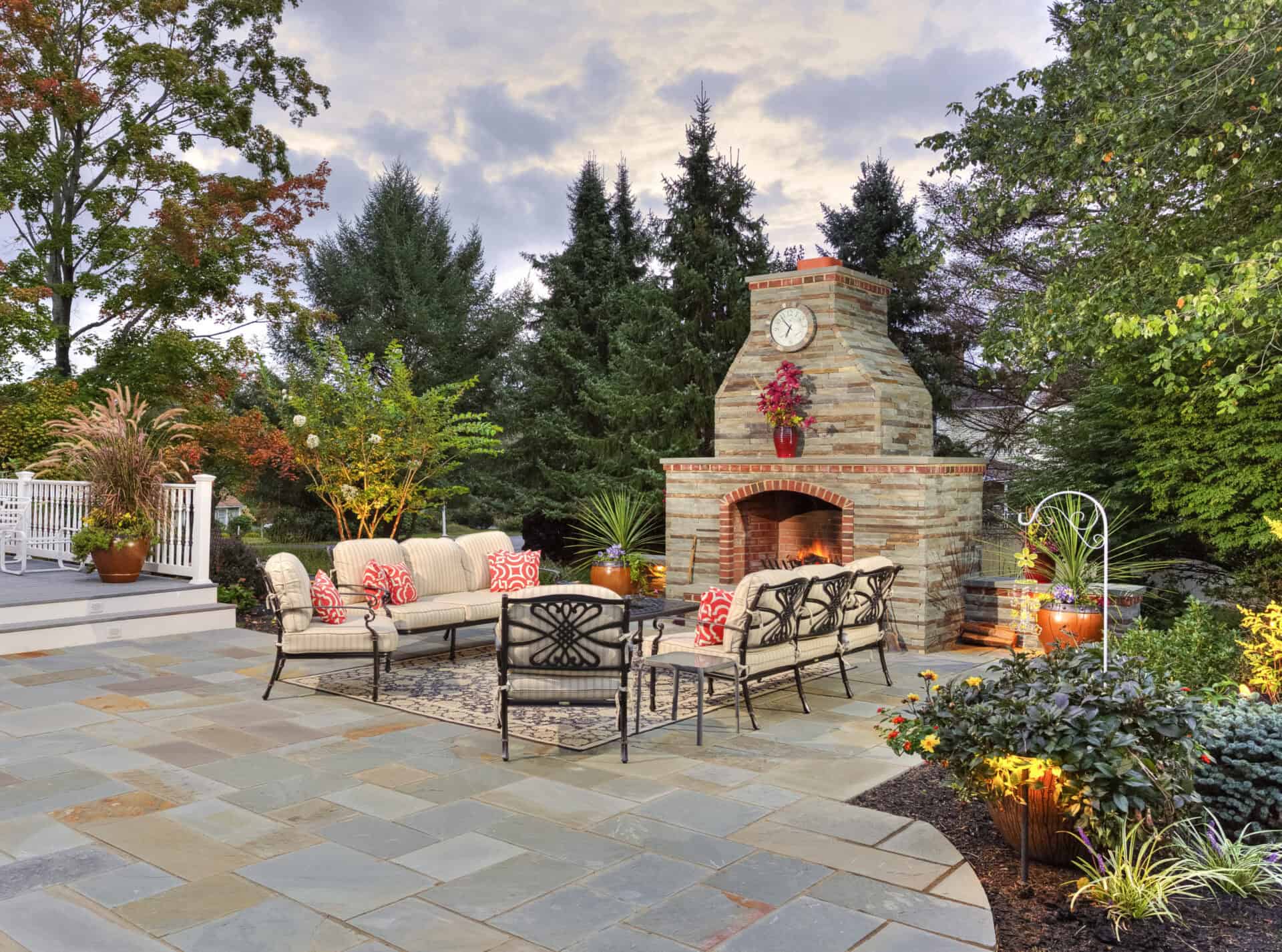 A spacious patio with cushioned seating, a natural stone fireplace with a clock, potted plants, and lush landscaping, surrounded by trees and garden lights at dusk.