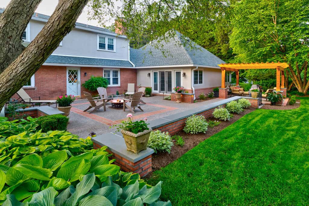 A backyard oasis with a brick patio showcasing Adirondack chairs around a fire pit. A pergola graces the space, surrounded by lush greenery, adjacent to a two-story house with a gray roof and red brick accents—a perfect example of outdoor living in Delaware County, PA.