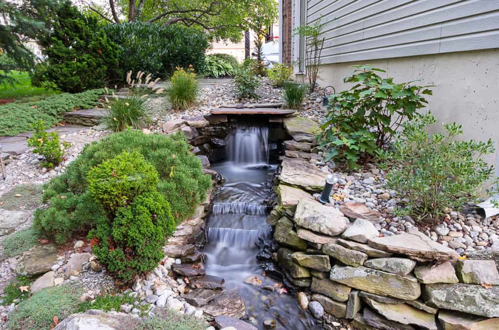 A small, tranquil garden waterfall flows over rocks, surrounded by lush greenery and shrubs. The water cascades into a stone-lined stream, crafting a peaceful setting beside a house with a beige exterior wall. For such idyllic outdoor living, consider landscaping services in Delaware County, PA.