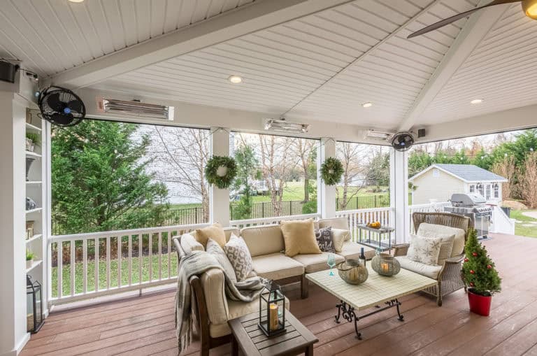 A spacious, covered outdoor patio with a white ceiling and wooden floor, featuring a cozy seating area with beige cushions, a coffee table, hanging wreaths, ceiling fans, and a small decorated Christmas tree. Expertly crafted by outdoor living specialists in Delaware County PA. A backyard and shed are visible in the background.