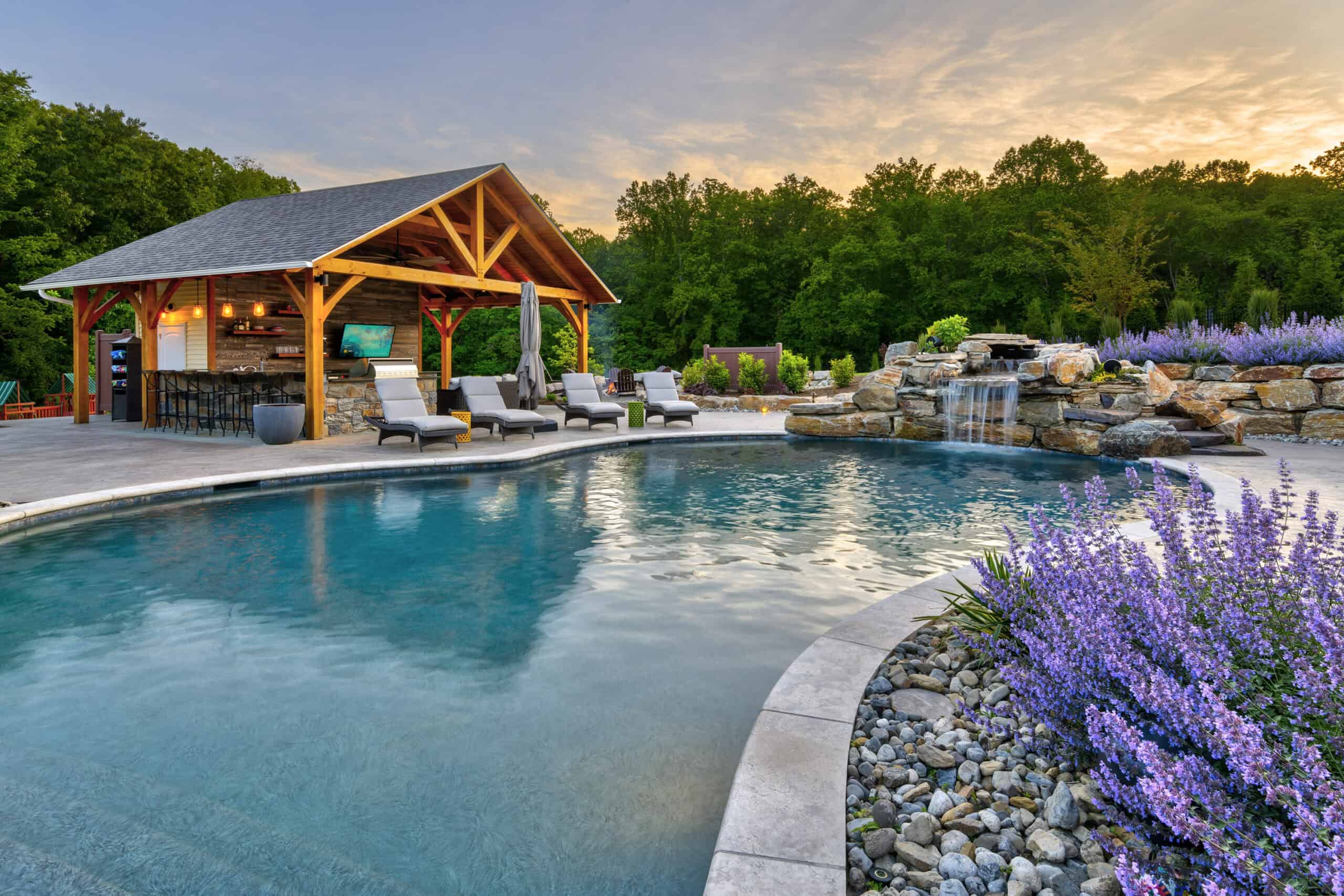 Luxurious backyard with a curved pool featuring a rock waterfall. Lounge chairs sit by the pool edge, next to a wooden gazebo housing a bar. Lush greenery and purple flowers line the area under a clear, golden sky—perfect for elegant outdoor living in Delaware County, PA.