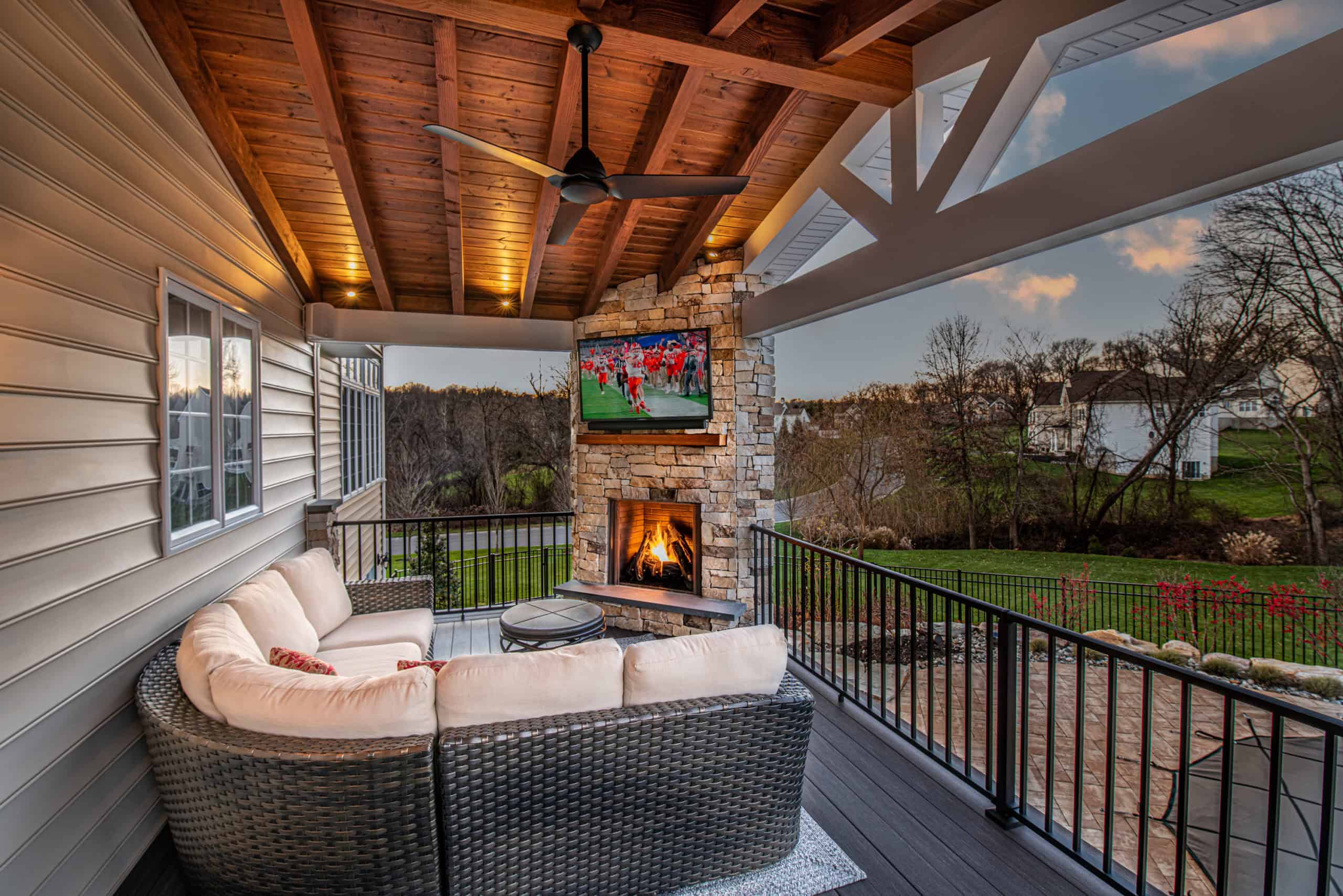 A covered outdoor patio with a wicker sectional sofa, a stone fireplace, a TV mounted above the fireplace, and a view of a green lawn and trees in the background at sunset.