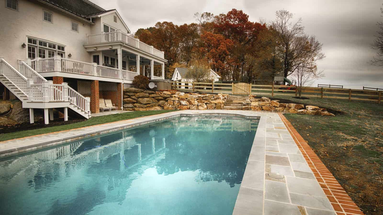 A backyard showcases a rectangular swimming pool with clear water, surrounded by stone and brick. A white, two-story house with a balcony overlooks the pool. In the background, autumn trees and a wooden fence enhance this serene outdoor living space in Delaware County, PA.