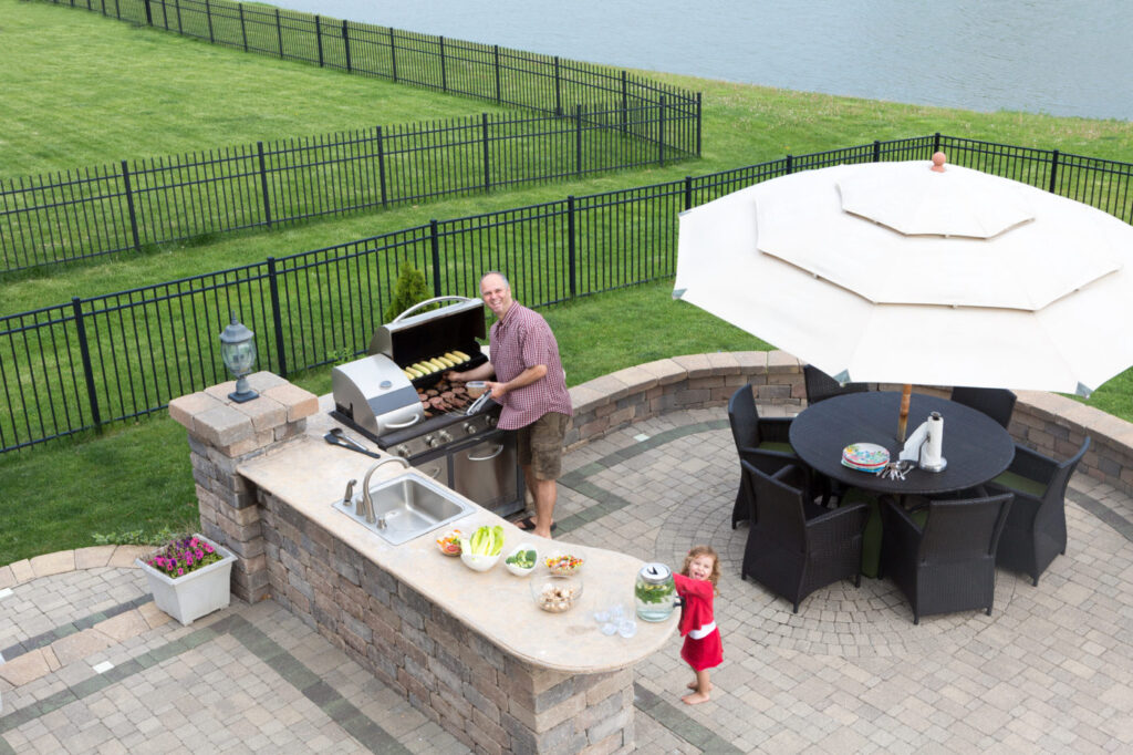 Father and daughter preparing a barbecue at an outdoor summer kitchen on a paved patio with a garden umbrella, table and chairs as they grill the meat on the gas BBQ waiting for guests to arrive