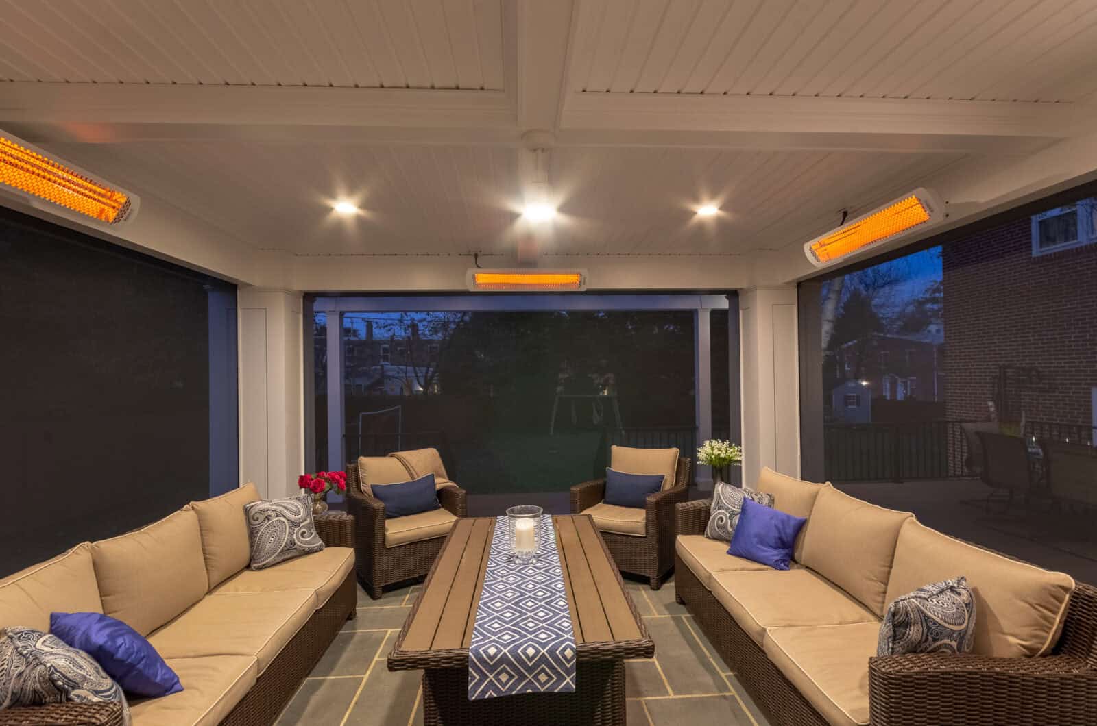 A cozy screened-in patio at dusk with wicker sofas, blue and beige cushions, a patterned runner on the coffee table, and overhead heaters lighting the space. A backyard and dark sky are visible outside.