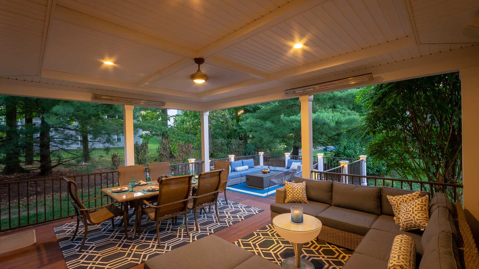 A cozy outdoor living area with cushioned seating, a dining table, and patterned rugs under a covered patio. Trees embrace the space, enhanced by warm lighting. Outdoor living in Delaware County, PA never felt more inviting with additional seating visible on the lower deck.