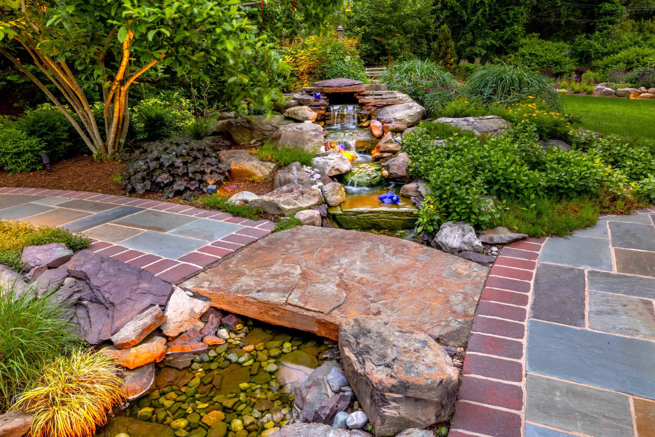 A landscaped garden features a small waterfall cascading over rocks into a pond. Surrounding greenery includes various plants and trees. The foreground showcases a colorful stone path bordered by bricks, a testament to expert landscaping services in Delaware County, PA, enhancing the serene natural scene.