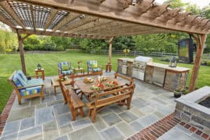 Wooden pergola with outdoor seating set on a stone patio. Features wooden tables and chairs with colorful cushions. A built-in stone kitchen area with a grill and a TV are in the background. Expertly crafted by outdoor living specialists in Delaware County, PA, it’s surrounded by lush green lawn and trees.