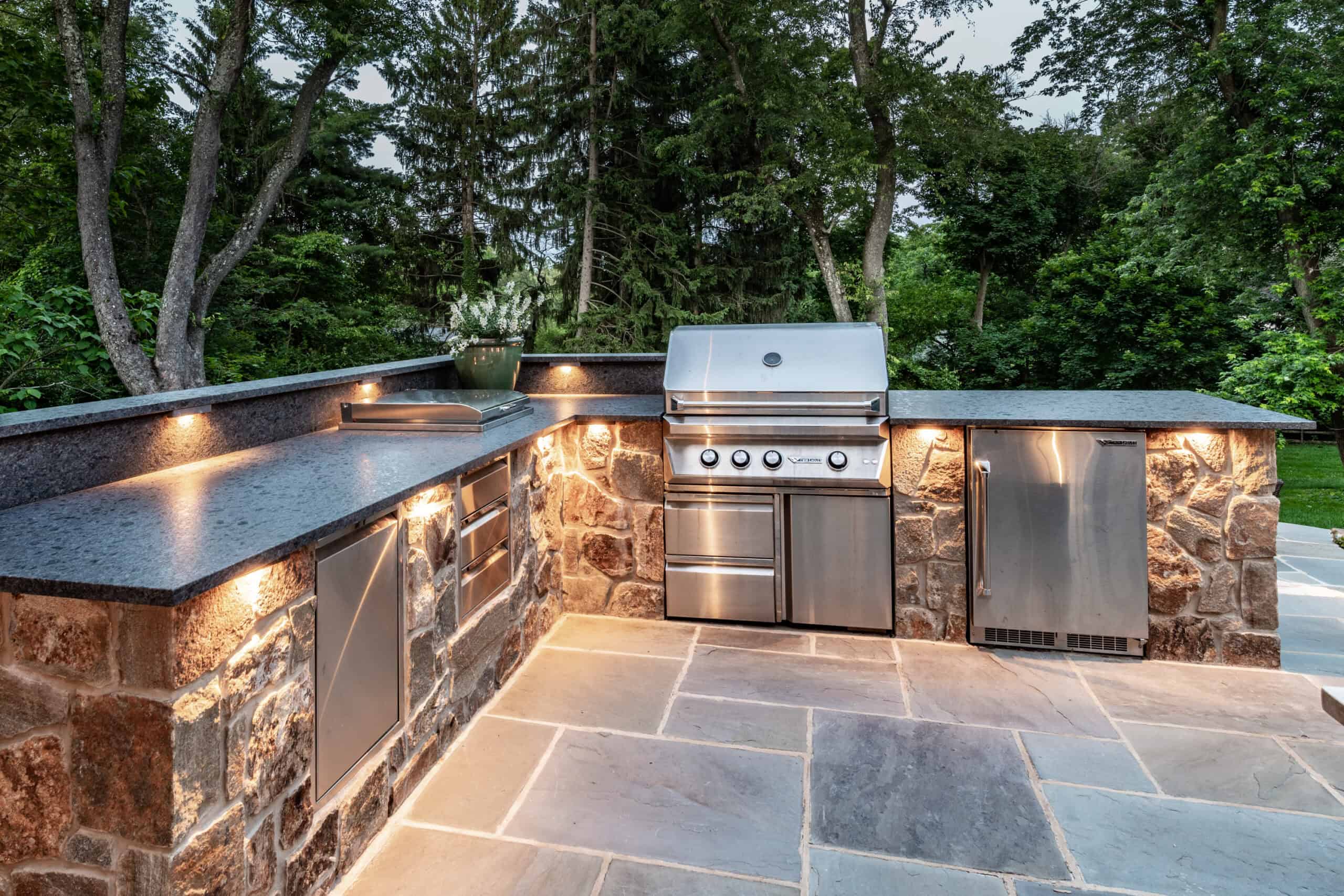 Modern outdoor kitchen with stone counters, stainless steel grill, fridge, and storage, set on a stone patio and surrounded by lush green trees; warm under-counter lighting creates an inviting atmosphere.