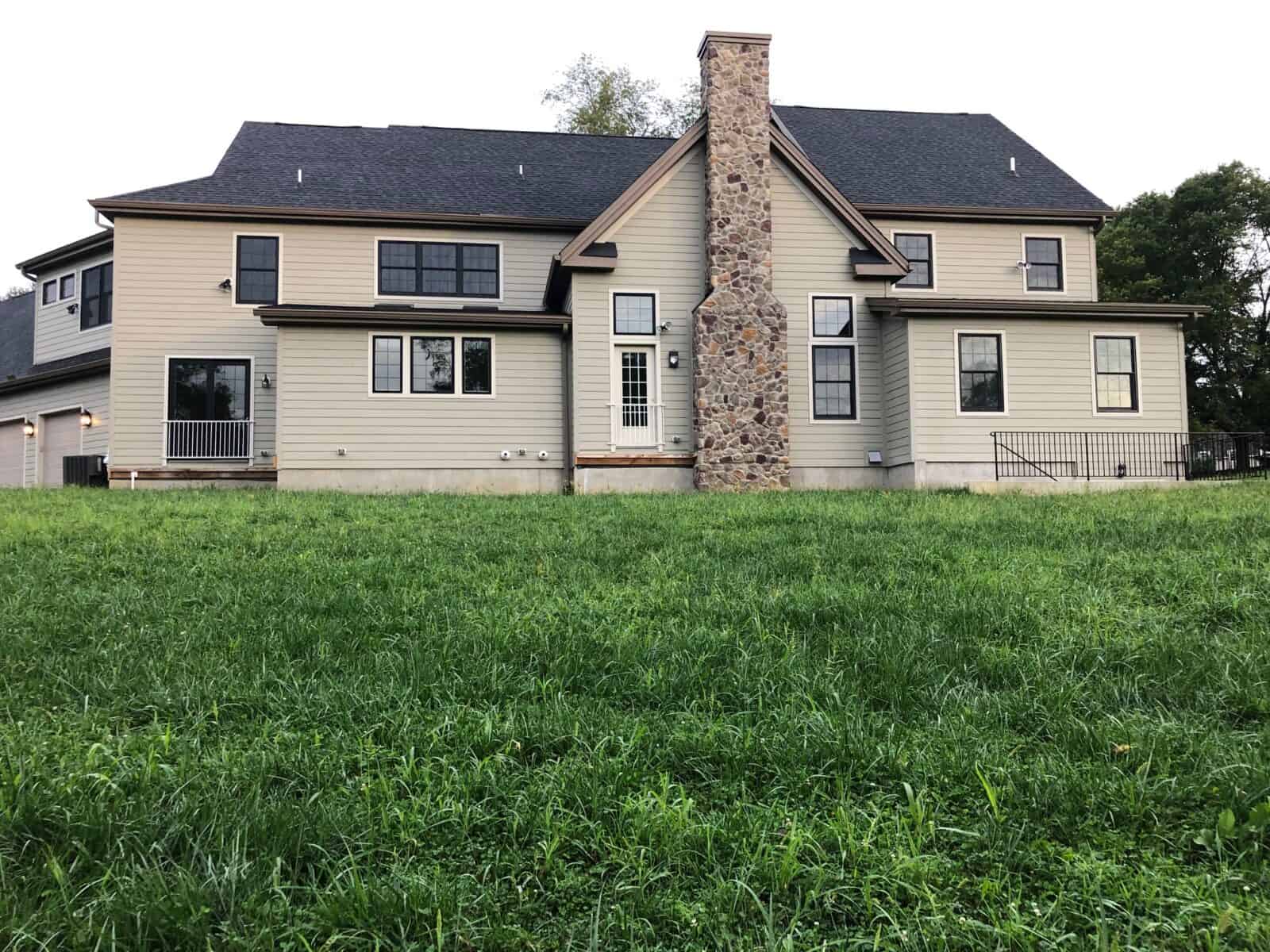 A two-story house with beige siding and a tall stone chimney graces a grassy lawn, showcasing outdoor living at its finest in Delaware County, PA. Numerous windows, a sloping roof, and a small side porch with railing complete the scene, framed by trees in the background.