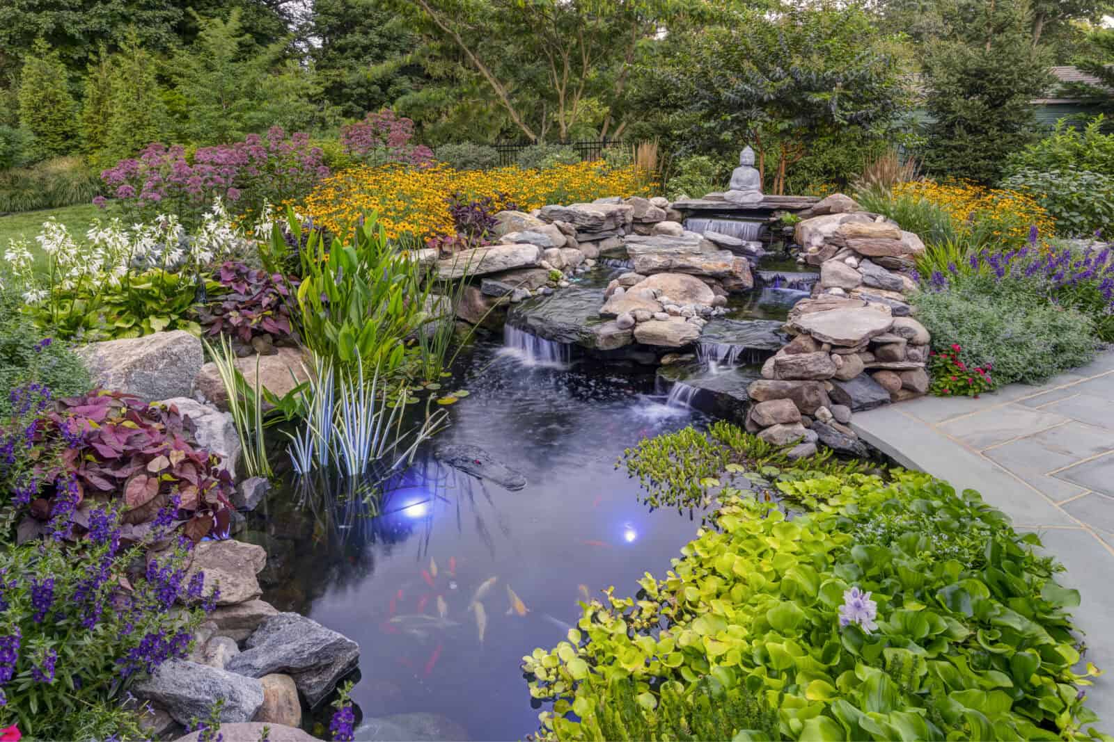 A serene garden in Delaware County, PA, features a koi pond with clear water and cascading rock waterfalls. Surrounding the pond are vibrant flowers, plants, and a small statue perched on rocks. Expert landscaping services ensure soft lights illuminate beneath the water for perfect outdoor living.