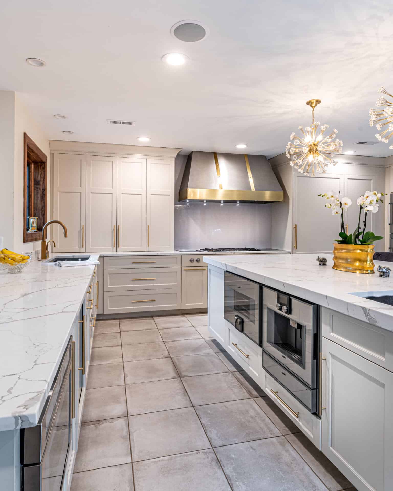 A modern kitchen featuring white cabinets, a marble countertop, a large stainless steel range hood, and a central island with built-in appliances. A chandelier hangs from the ceiling, and a potted orchid decorates the island—a perfect indoor complement to your outdoor living space in Delaware County PA.