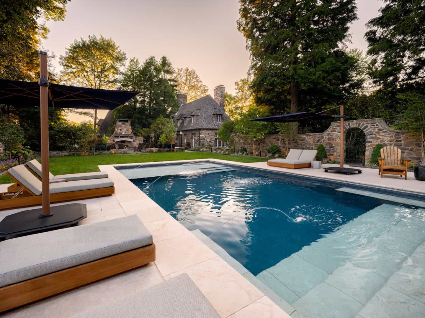 A serene backyard in Delaware County, PA, features a rectangular swimming pool surrounded by expertly designed hardscaping and lounge chairs. A stone house is visible in the background, framed by trees and a stone wall, all bathed in a soft evening glow perfect for outdoor living.
