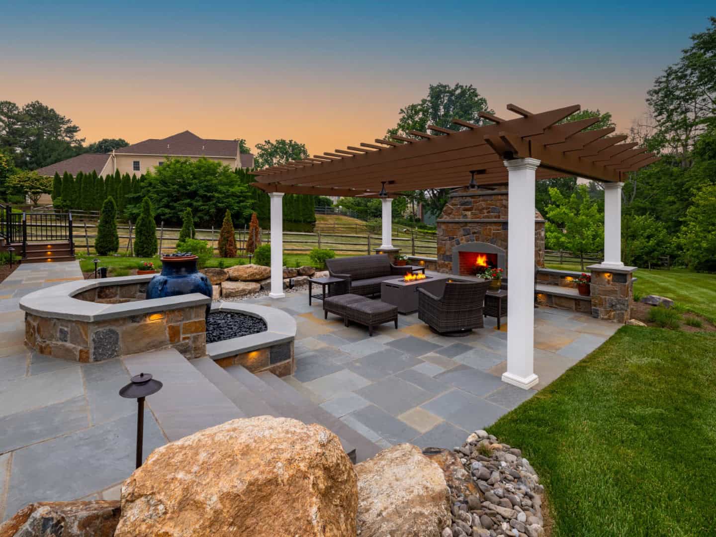 Luxurious outdoor patio at sunset with a stone fire pit and comfortable wicker chairs under a large pergola. The lush setting is enhanced by expert hardscaping services in Delaware County, PA, boasting beautifully arranged rocks and hedges with a spacious lawn beyond.