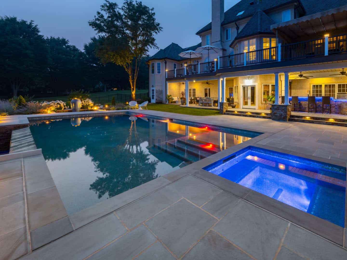 A luxurious backyard, crafted with expert landscaping services in Delaware County PA, features a large swimming pool and a hot tub illuminated at dusk. Trees and well-lit seating areas complement the spacious house with balconies and large windows, enhancing the serene atmosphere.
