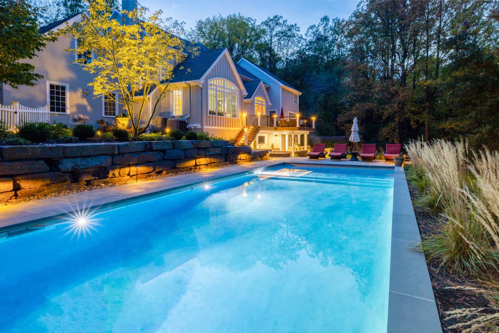 A well-lit, modern house with large windows is set against a backdrop of trees during dusk. A rectangular swimming pool with glowing lights reflects the house, showcasing exquisite outdoor living in Delaware County, PA. Red lounge chairs and a large umbrella are by the poolside.