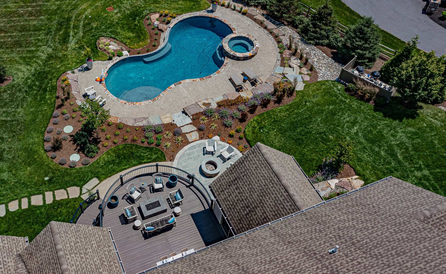 Aerial view of a landscaped backyard in Delaware County, PA, showcasing a curvy swimming pool with a hot tub. Adjacent is a circular stone fire pit surrounded by furniture on a wooden deck. This outdoor living space features well-manicured lawns and pathways, perfect for relaxation.