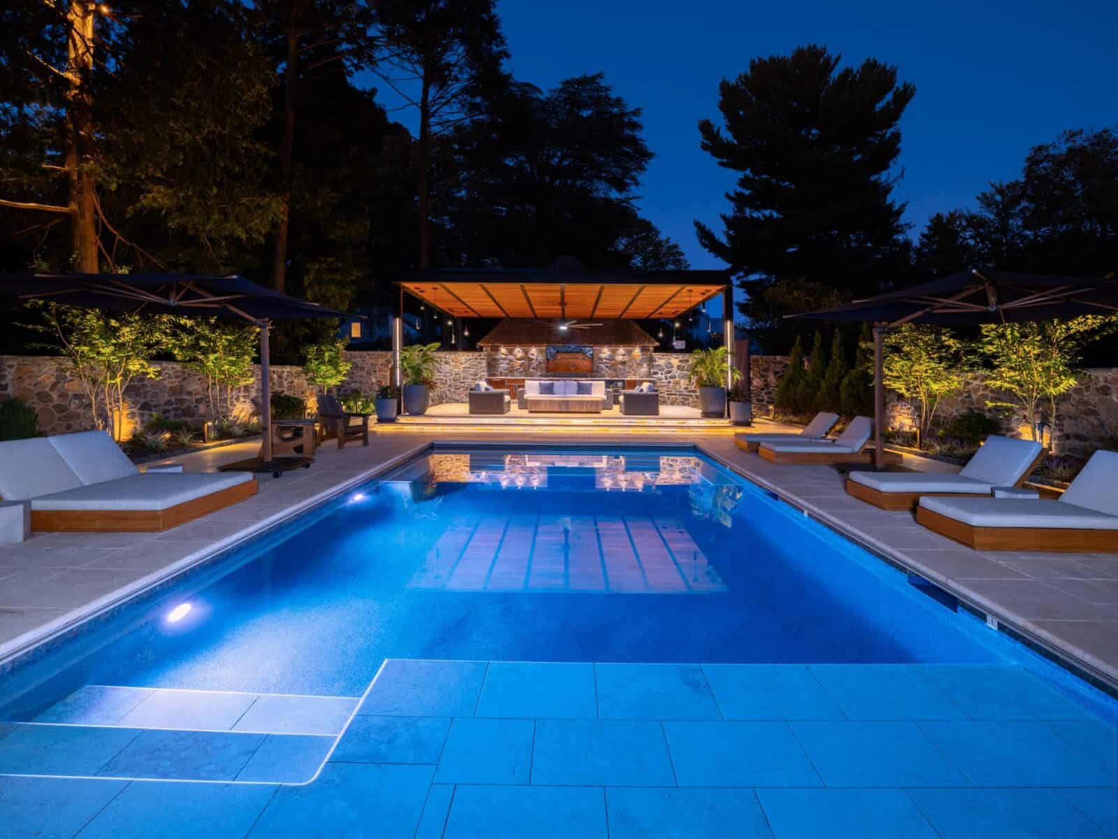 A luxurious outdoor pool area at twilight, surrounded by cushioned loungers and shaded pergolas. Expert hardscaping services in Delaware County, PA ensure the pool is illuminated beautifully, reflecting the ambient lighting. Trees and stone walls add privacy to this serene sanctuary.