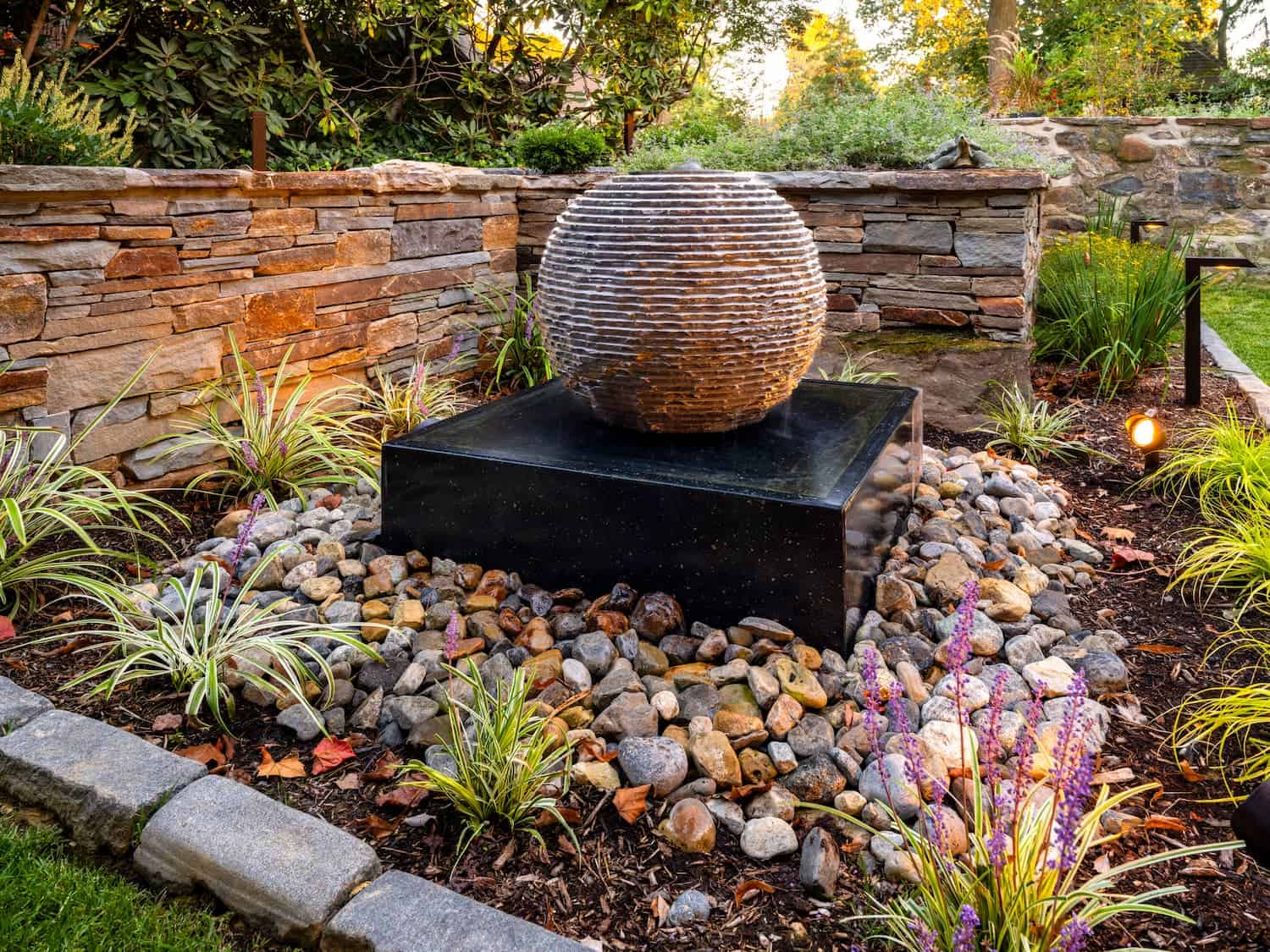 A stone sphere fountain sits on a black pedestal, surrounded by river rocks and ornamental grasses, in a landscaped garden bordered by a stone wall. Warm lights highlight the peaceful outdoor setting.