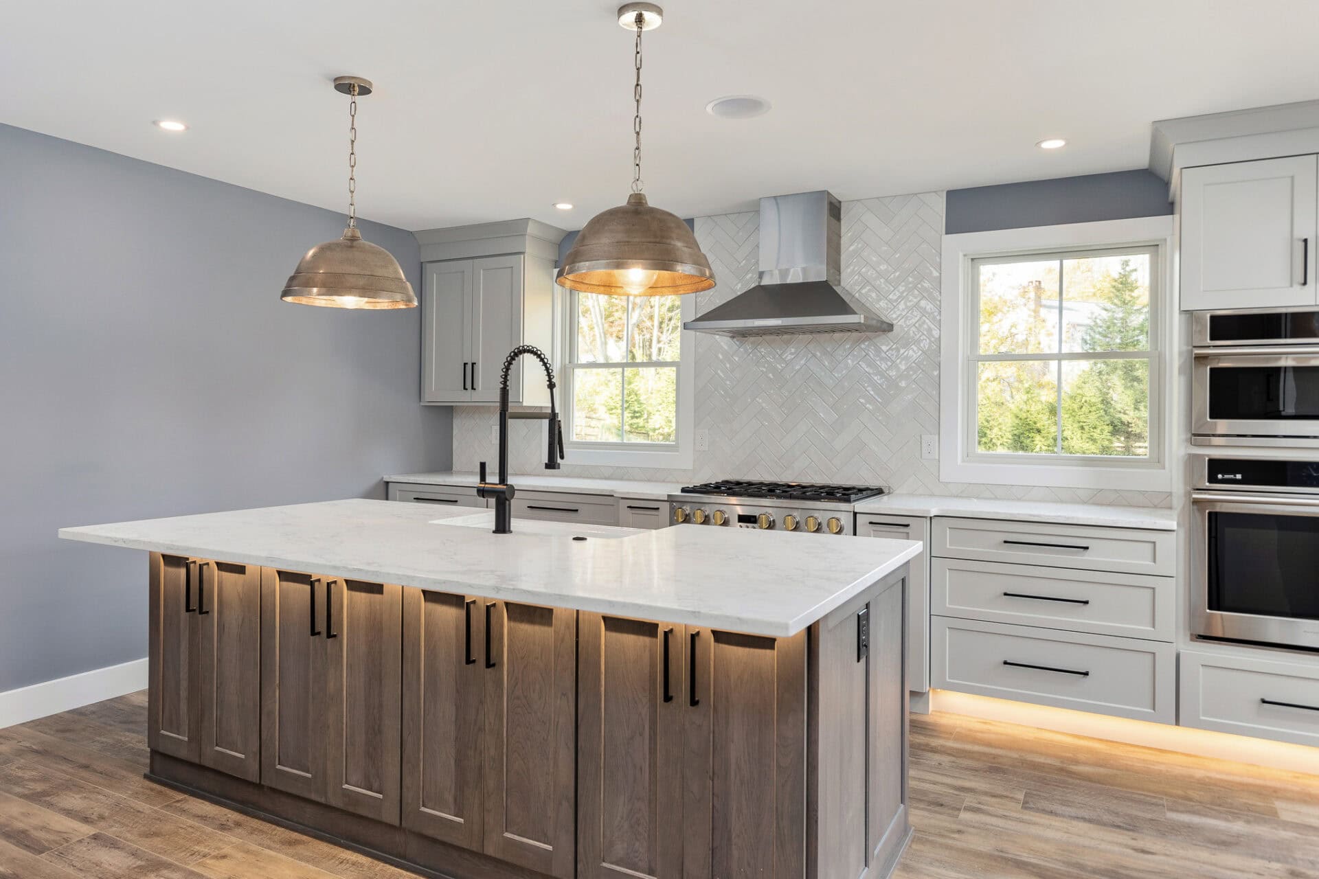 Modern kitchen with a large island featuring dark wood cabinets and a white marble countertop. Two pendant lights hang above. Gray cabinets, stainless steel appliances, and a herringbone backsplash complete the space, blending indoor elegance seamlessly with outdoor living in Delaware County PA.