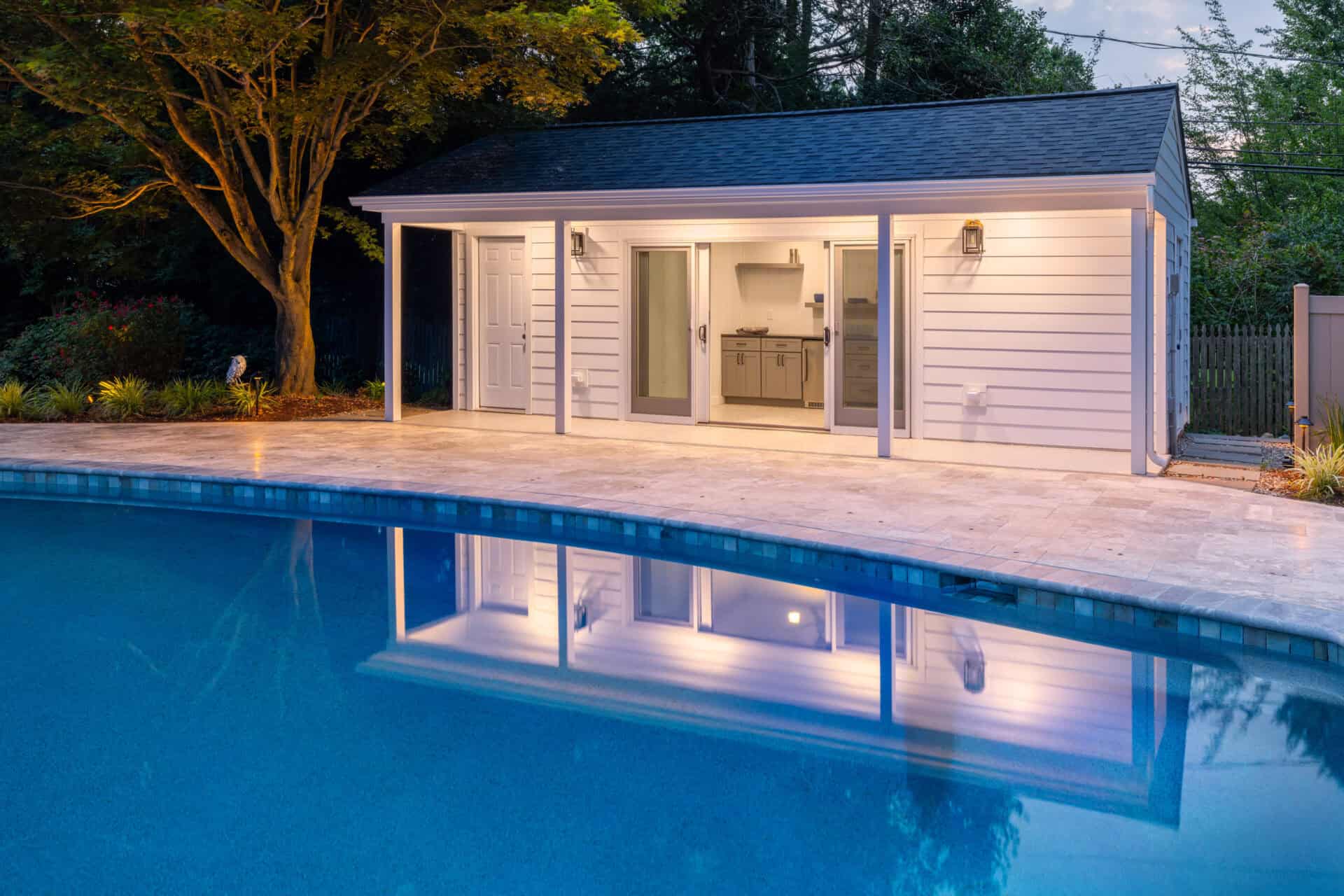A modern pool house with white siding and large glass doors is illuminated at dusk, epitomizing outdoor living in Delaware County, PA. It stands beside a tranquil pool with reflective water, bordered by expertly crafted stone paving. Tall trees and a fence are visible in the background.