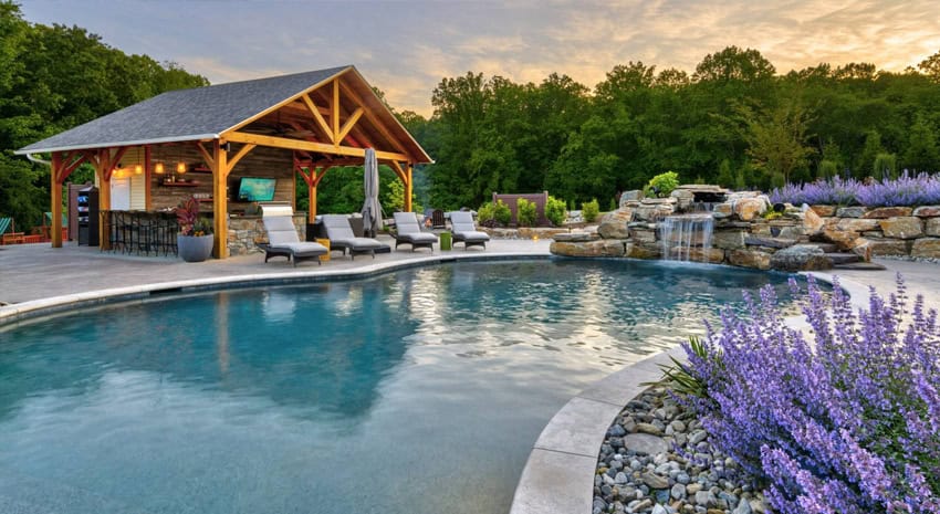 A luxurious outdoor pool with lounge chairs, a wooden pavilion bar, and a stone waterfall feature surrounded by purple flowers and greenery at sunset.