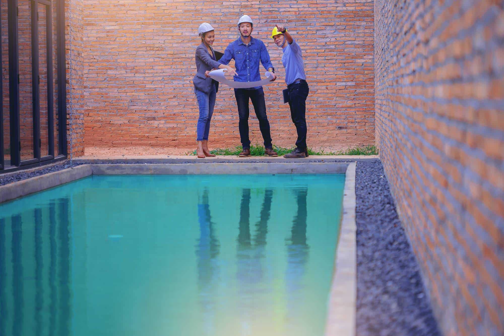 Three people in construction helmets stand by a swimming pool, examining blueprints. One person points forward, and two brick walls border the pool area. The setting appears to be a construction or inspection site.