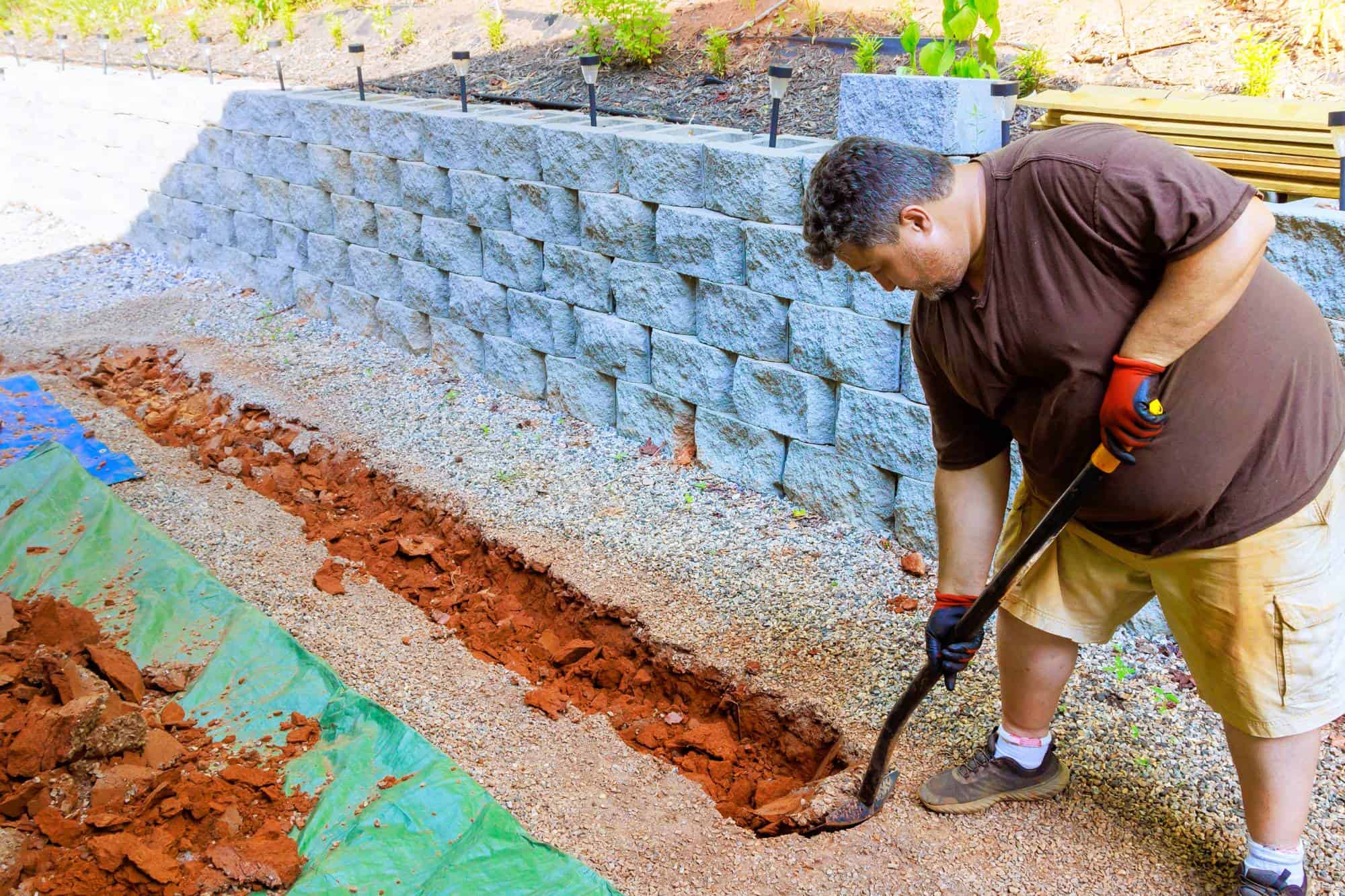 A man wearing gloves uses a shovel to dig a trench in reddish soil beside a stone retaining wall in a backyard, with green tarps and gravel on the ground.