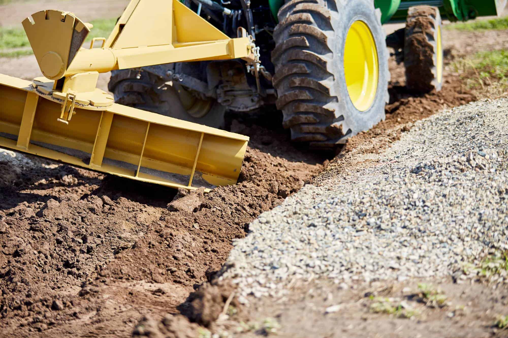 A close-up of a tractor with a yellow grader attachment leveling soil next to a gravel path, preparing the ground for construction or landscaping work.