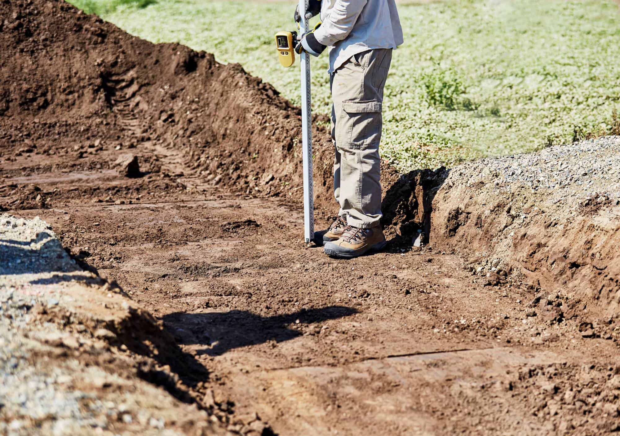A person in workwear and boots uses a measuring tool to check ground level on a dirt construction site, with soil and gravel visible around them.