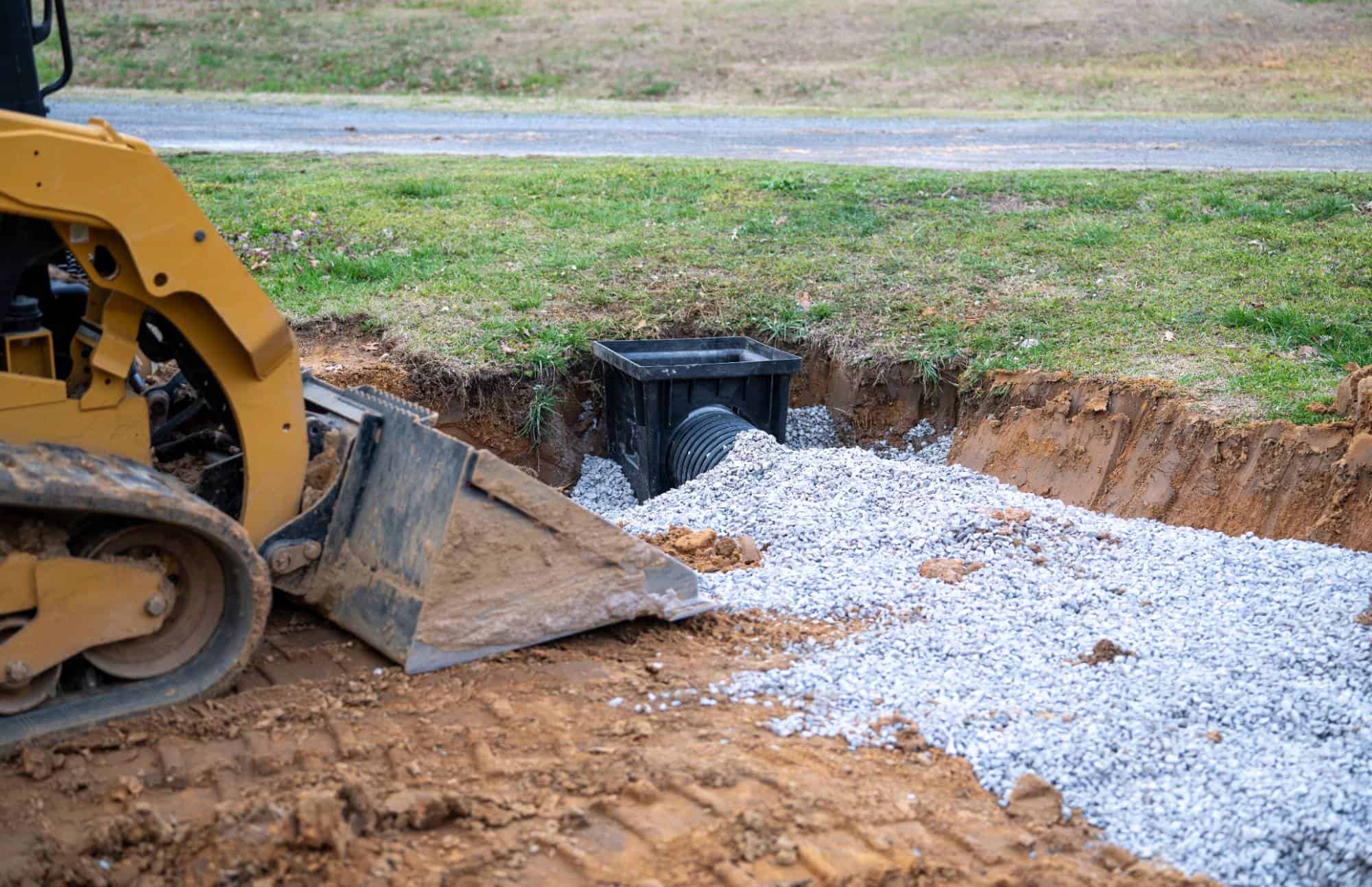 A skid steer loader dumps gravel into a trench leading to a black drainage box with a large pipe, set in a grassy area near a road.