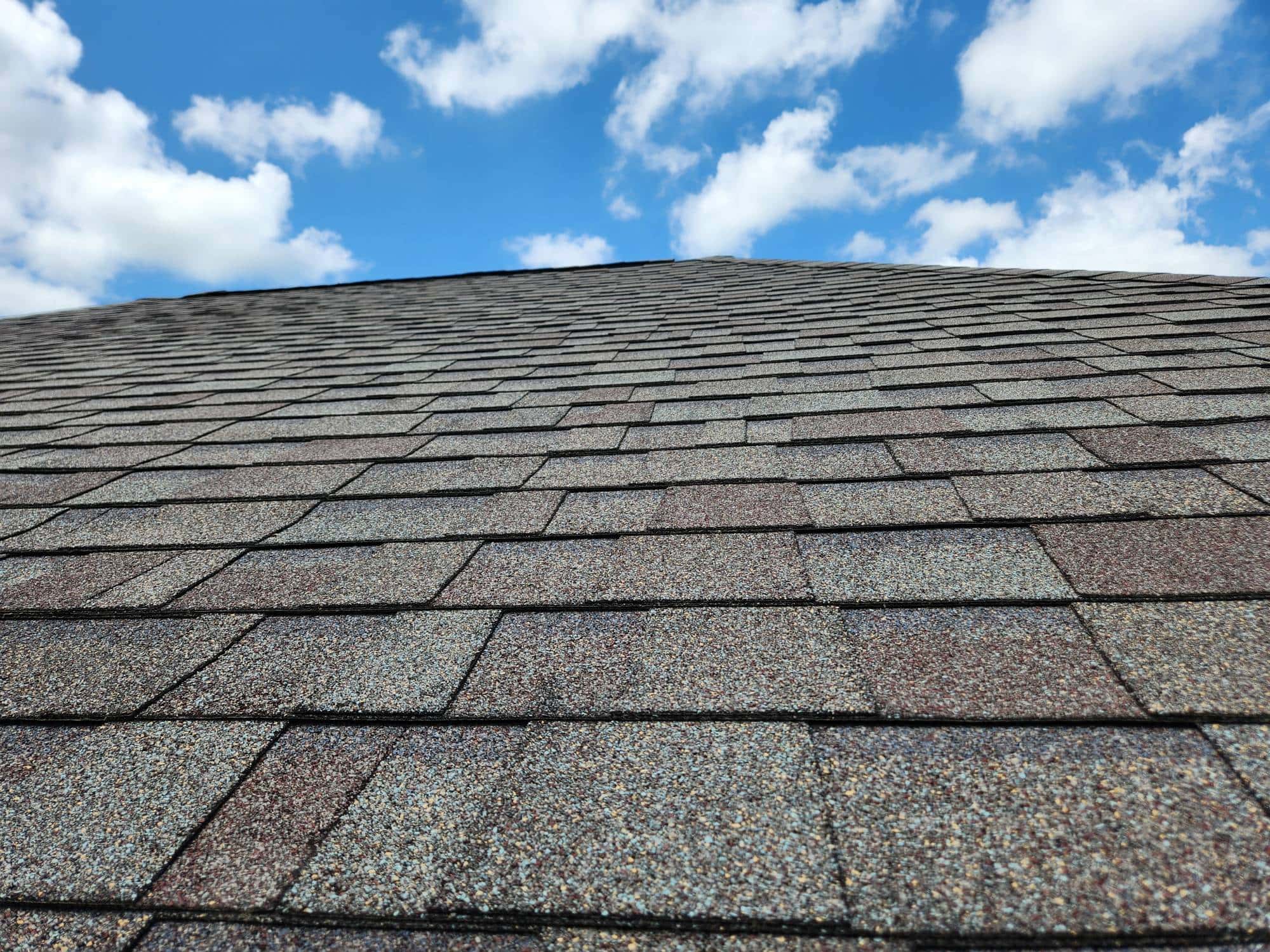 The image shows a close-up view of a rooftop covered with asphalt shingles, stretching upward toward a bright blue sky with scattered white clouds.